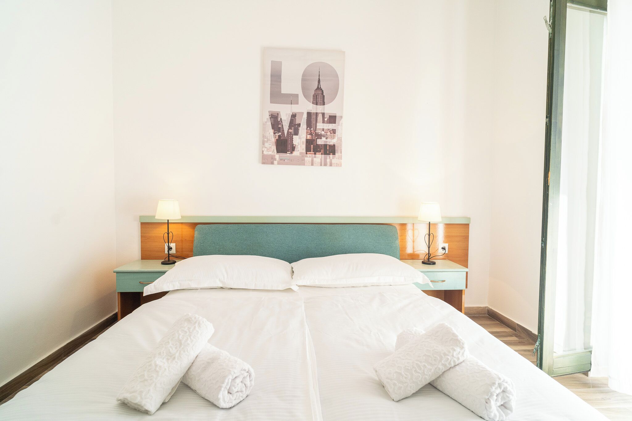 Une chambre d’hôtel avec un lit, deux tables de chevet avec des lampes, une tête de lit et un mur orné d’un panneau indiquant le mot « AMOUR ».