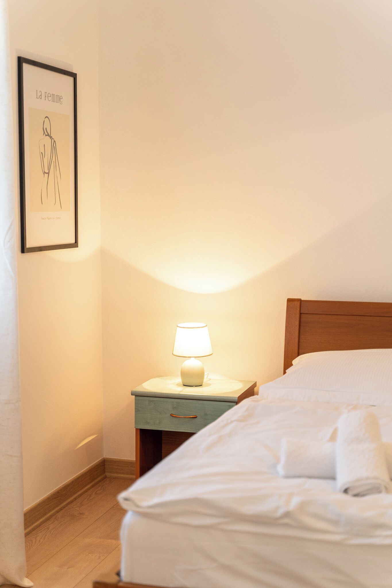 Un lit bien fait, avec du linge de lit blanc, une table de chevet avec une lampe et un tableau encadré au mur.