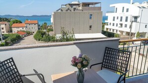 Een balkon met een tafel en stoelen, een vaas met bloemen en uitzicht op zee en gebouwen.
