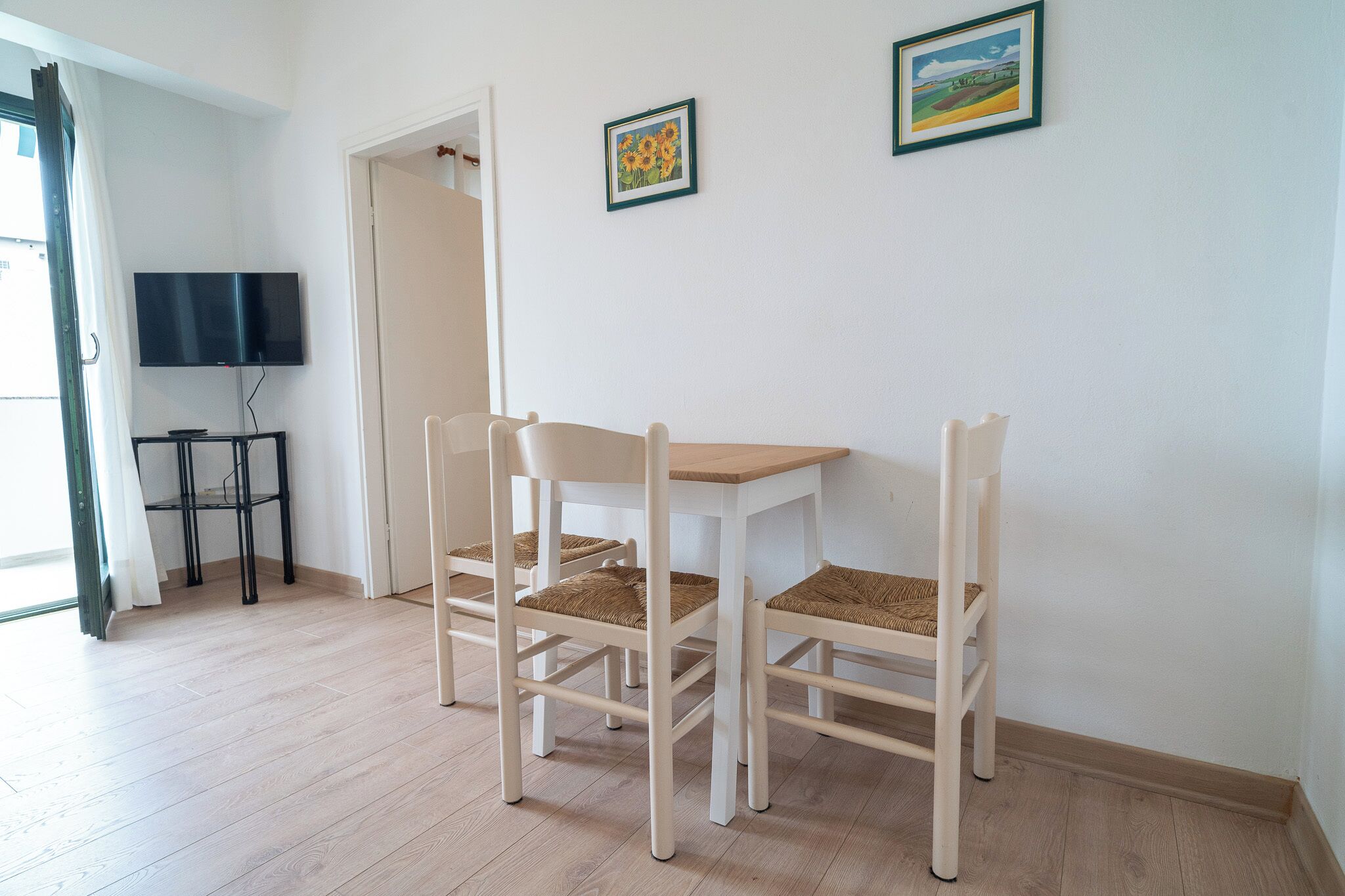Une pièce avec un plancher en bois, une table blanche avec deux chaises, un téléviseur posé sur un meuble et deux tableaux encadrés accrochés au mur.