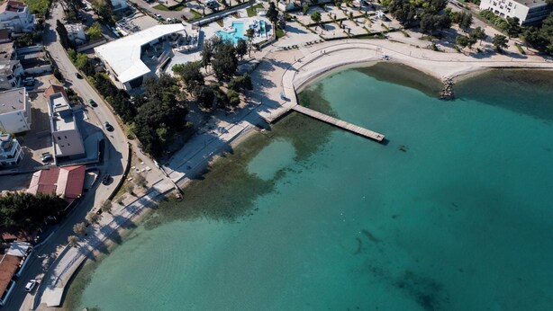 Luchtbeeld van een kustgebied met een pier die zich uitstrekt in turquoise water, omringd door gebouwen en een bochtige weg.