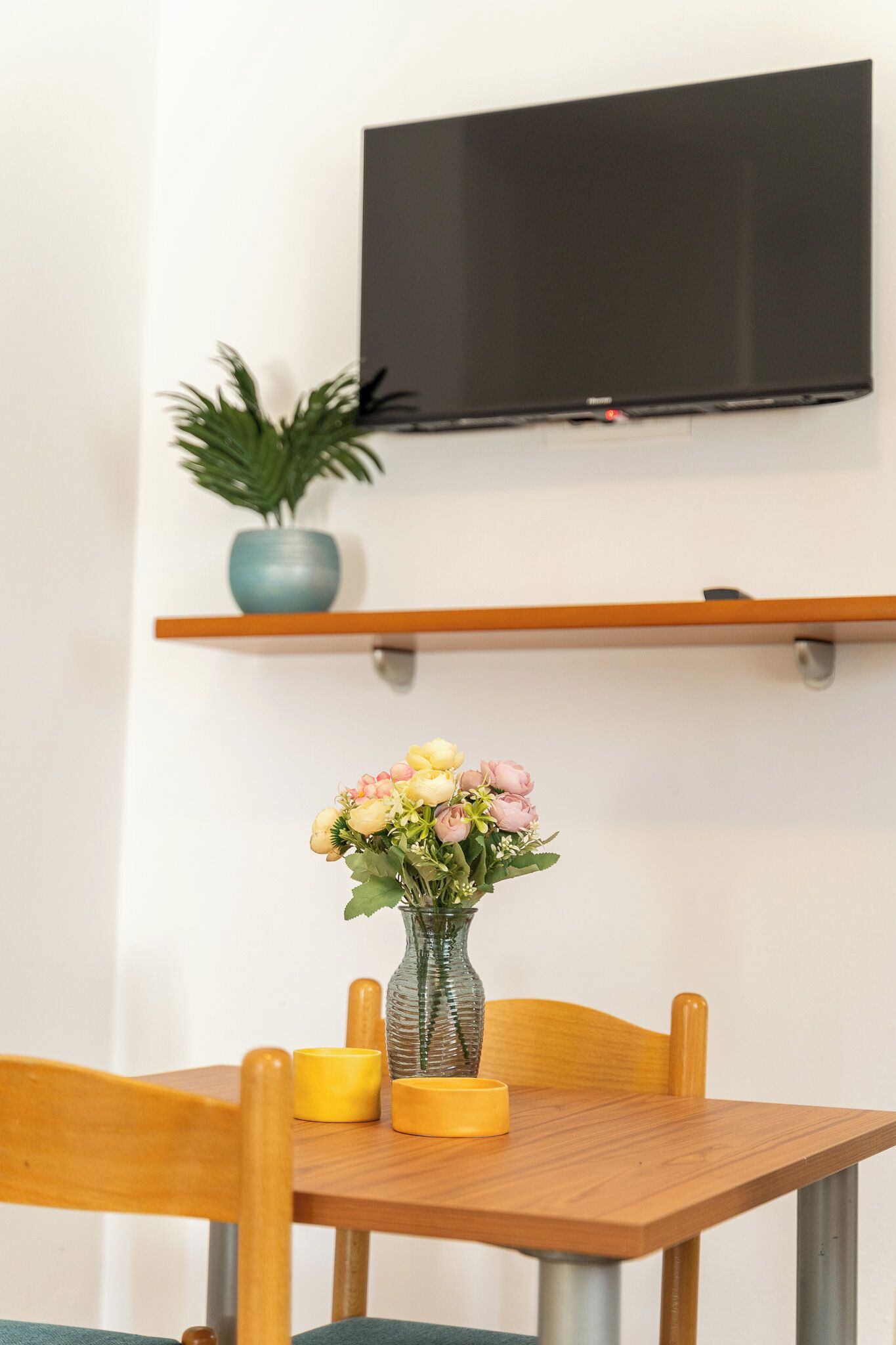 A dining table with a vase of flowers and candles, a television mounted on the wall, and a shelf with a plant.