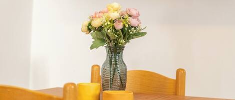 A dining table with a vase of flowers and candles, a television mounted on the wall, and a shelf with a plant.