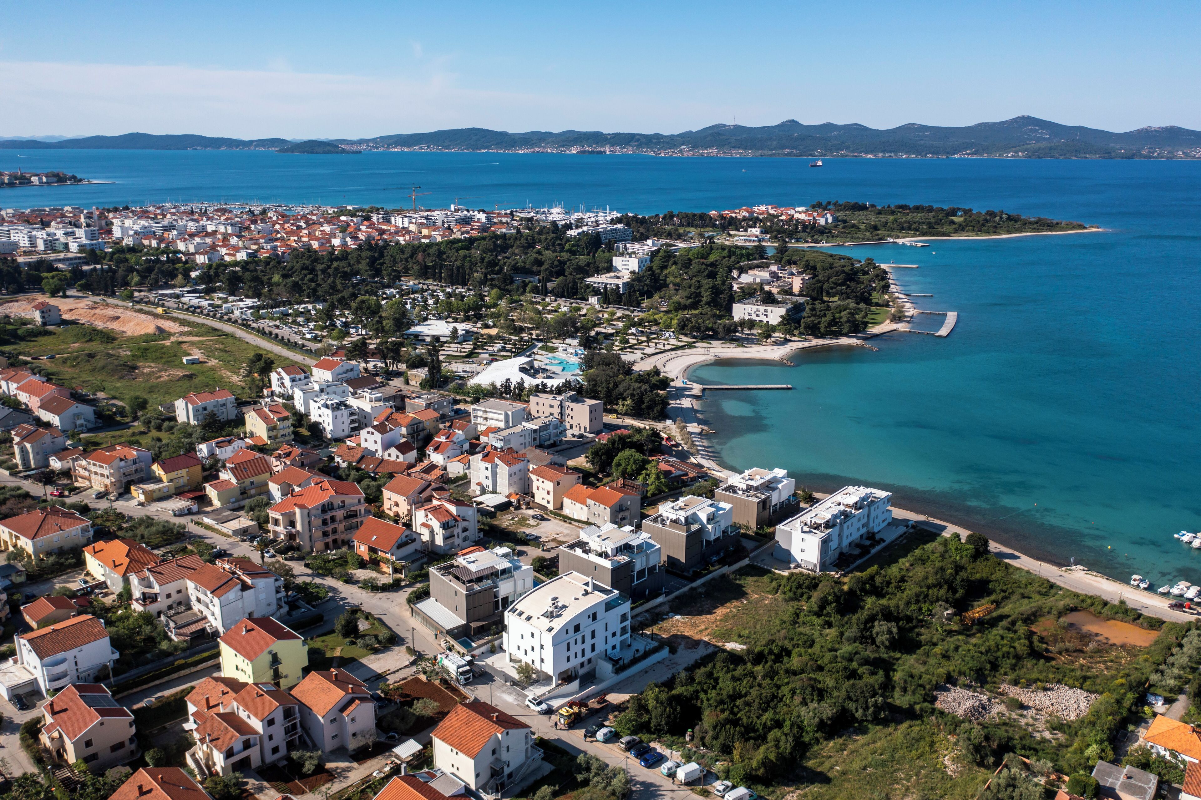 An aerial view of a coastal town with residential buildings, a beach, and a clear blue sea.