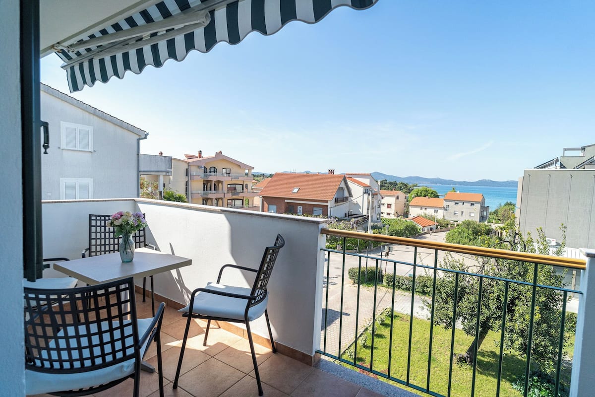 A balcony with a table and chairs, a view of the sea, and neighboring buildings.