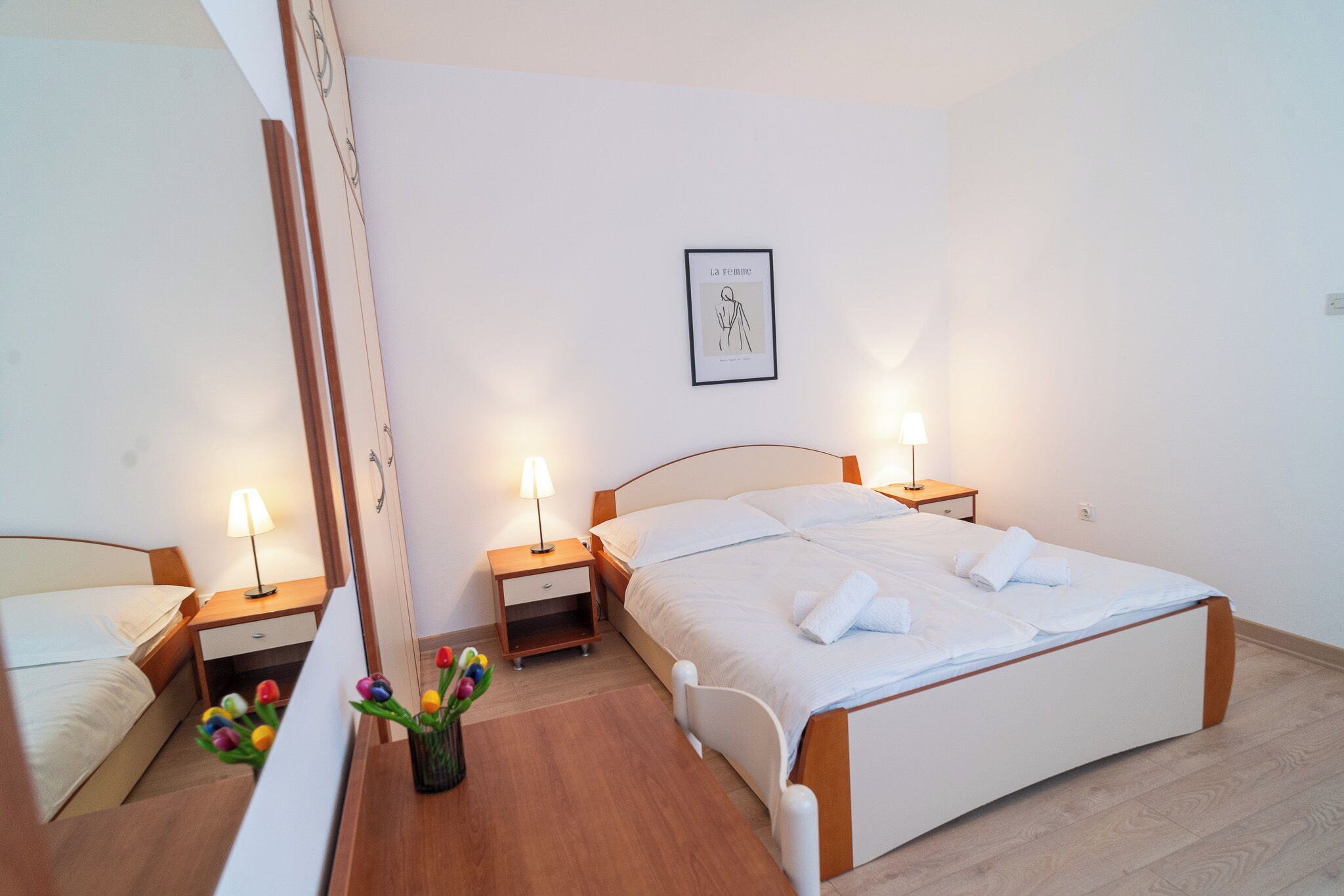 A neatly arranged bedroom with a large bed, two bedside tables with lamps, a bedside drawer, and a wall-mounted picture.