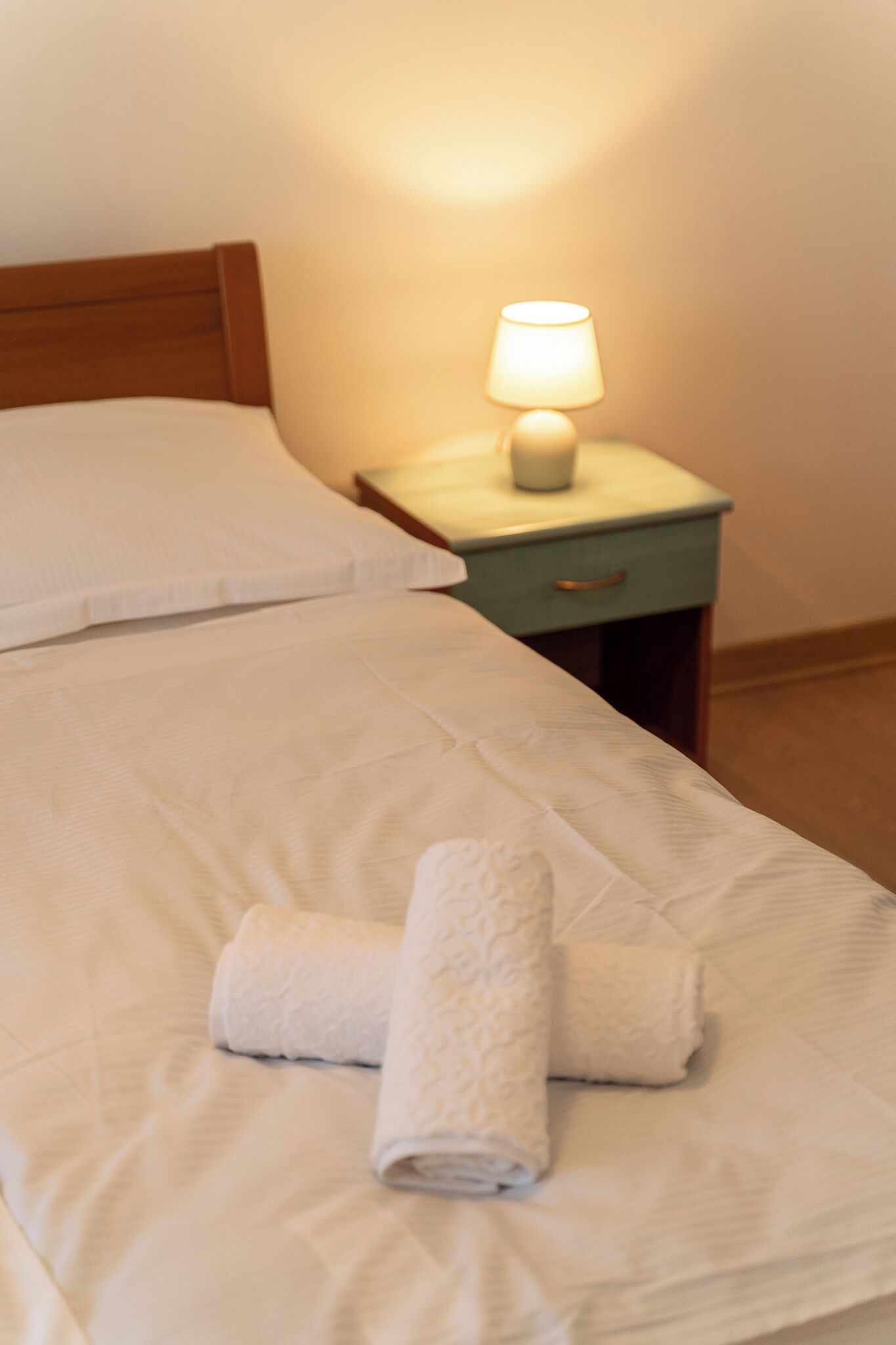 Een net opgemaakt bed met witte lakens en twee opgerolde handdoeken. Een nachtkastje met een lamp en een klein lade is zichtbaar.
