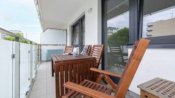 Un balcón con mesa y sillas de madera, un taburete pequeño y una ventana con vistas a edificios y árboles.