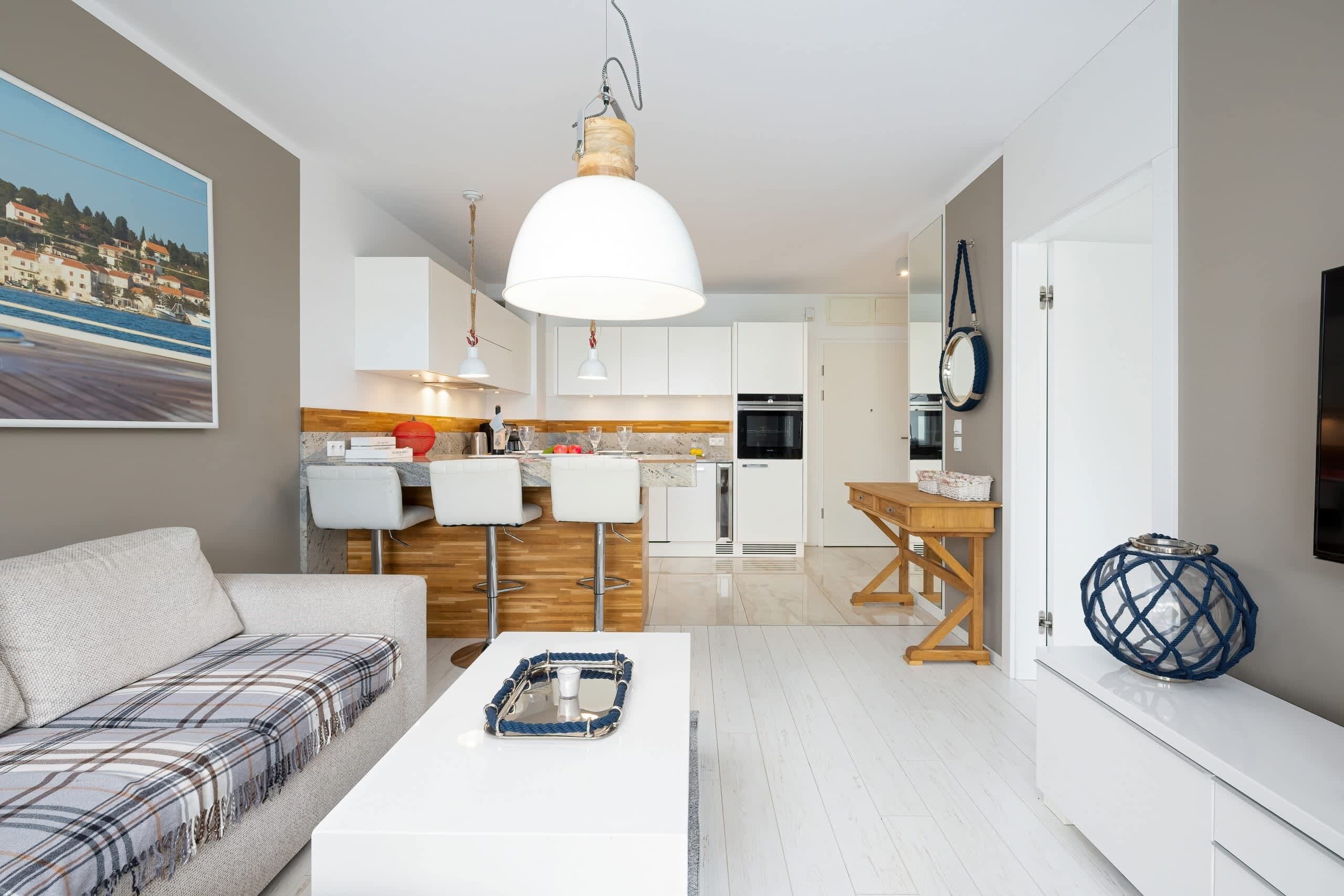 Una sala moderna con un sofá, una mesa de centro y una zona de cocina con armarios blancos y una lámpara colgante.