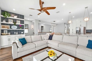 A modern living room with a sectional sofa, a coffee table with flowers, a ceiling fan, and a kitchen in the background.
