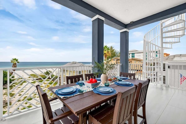 A dining area with a table set for four, overlooking a beach with palm trees and a clear sky.