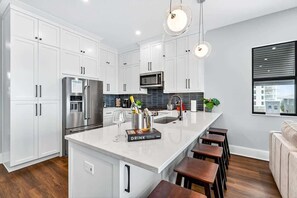 A modern kitchen with white cabinetry, stainless steel appliances, a central island with bar stools, and a large window with blinds.