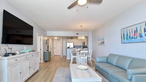 A modern living room with a flat-screen TV, a light blue sofa, a dining area with white chairs, and a kitchen in the background. - Condo in the heart of Destin! Lagoon-style pool and tons of great amenities! (Destin)