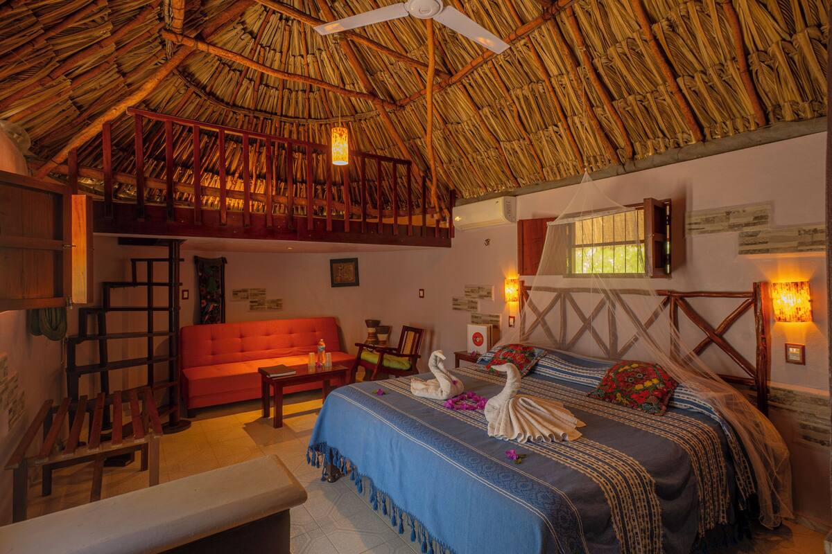 Una habitación con techo de paja, una cama con colcha azul, un sofá rojo y un banco de madera.