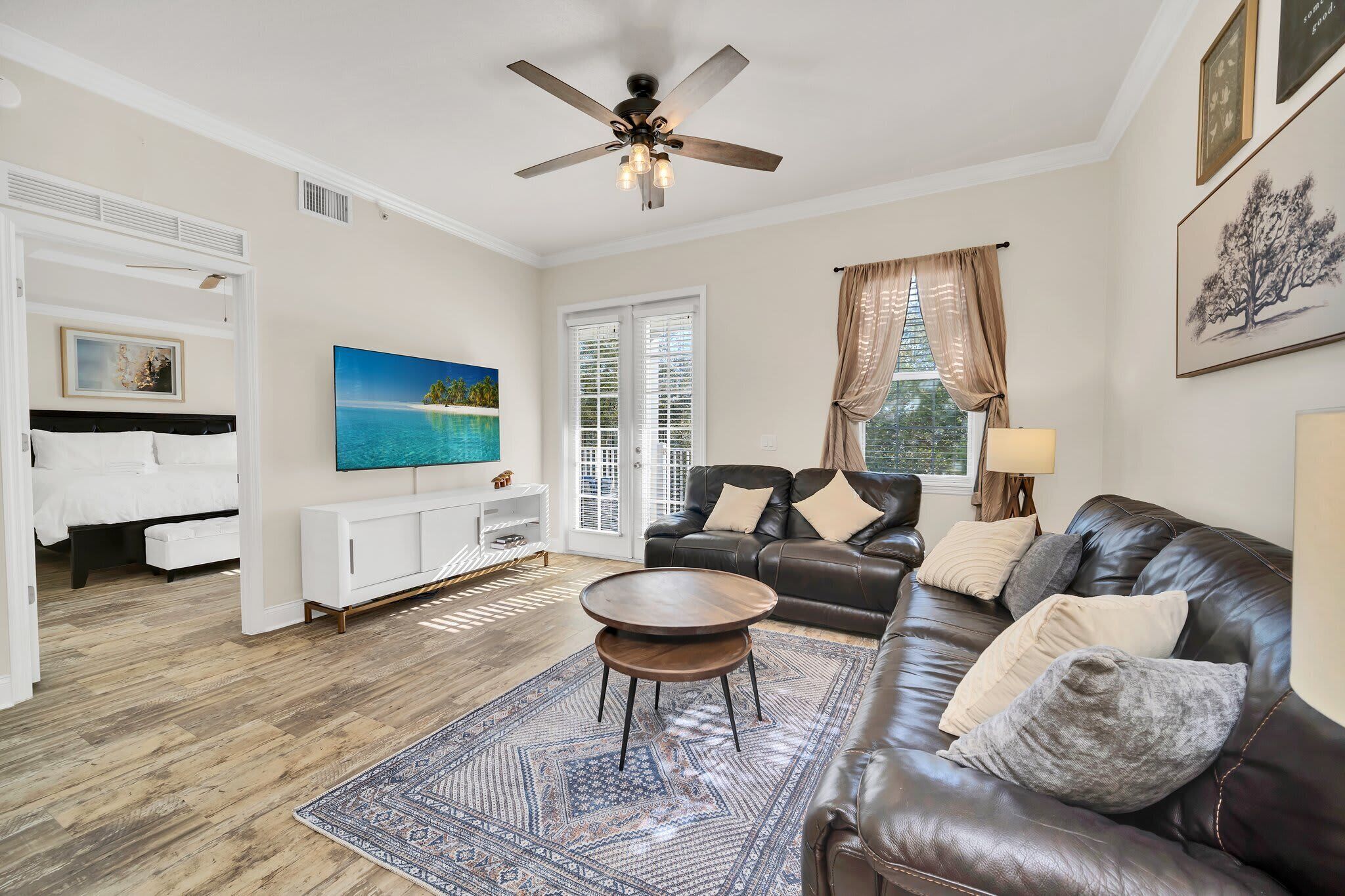 Un salon avec un canapé en cuir marron, une table basse ronde, un tapis à motifs, un ventilateur de plafond et un tableau représentant une plage.