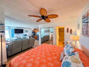 Un dormitorio con cama, ventilador de techo, baño con ducha y sala de estar con sofá y televisor.
