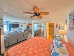 A bedroom with a bed, a ceiling fan, a view of the living area, and a door leading to a bathroom. - Estero Beach & Tennis Club #103C Beachfront Condo (Fort Myers Beach)
