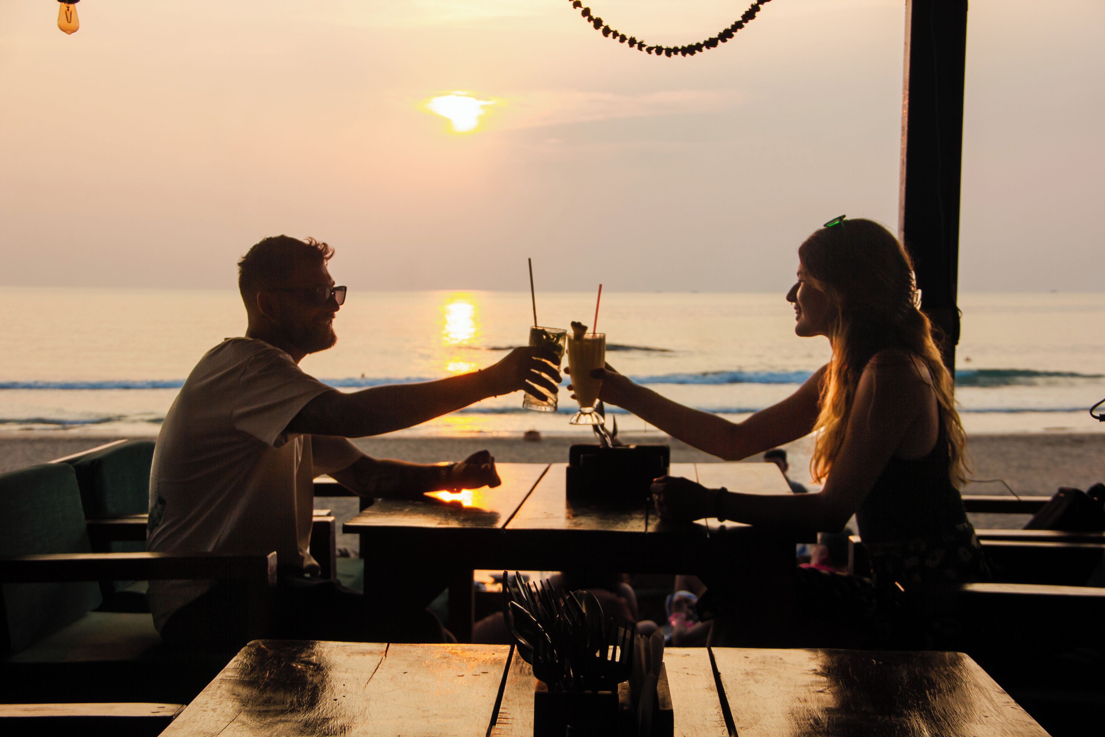 A couple toasting with drinks at a wooden table overlooking the ocean at sunset.