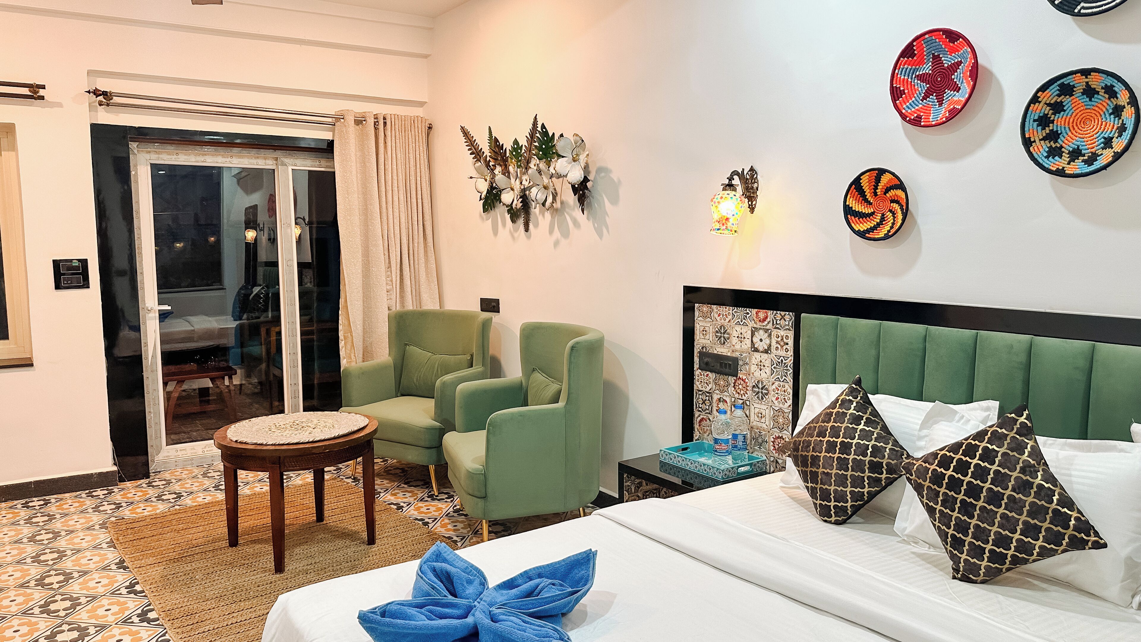Habitación de hotel con cama, dos sillones verdes, una mesita redonda, un adorno de pared y una pared estampada.