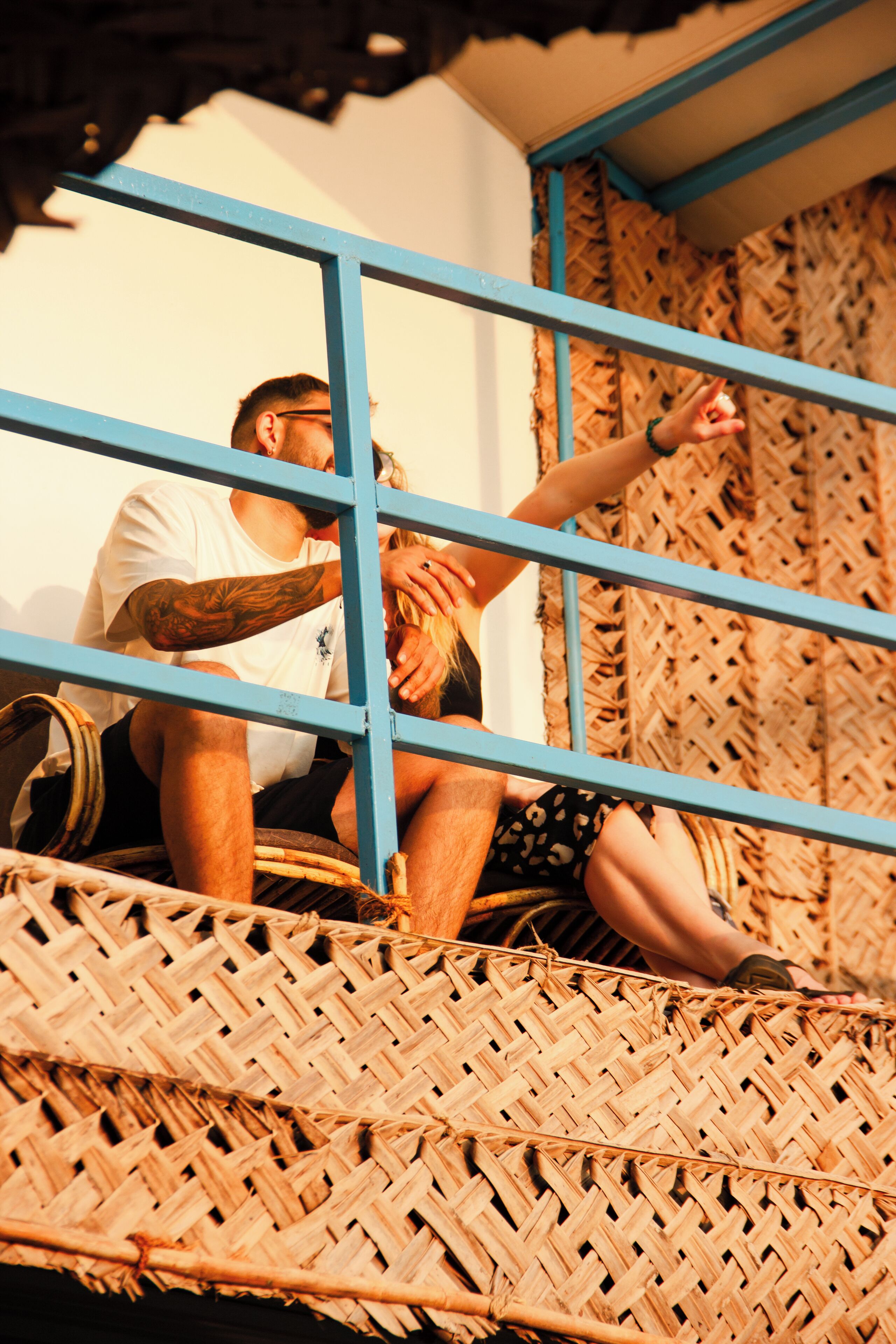 A person sitting on a woven bench with a metal railing, a woven wall behind, and a white wall to the side.