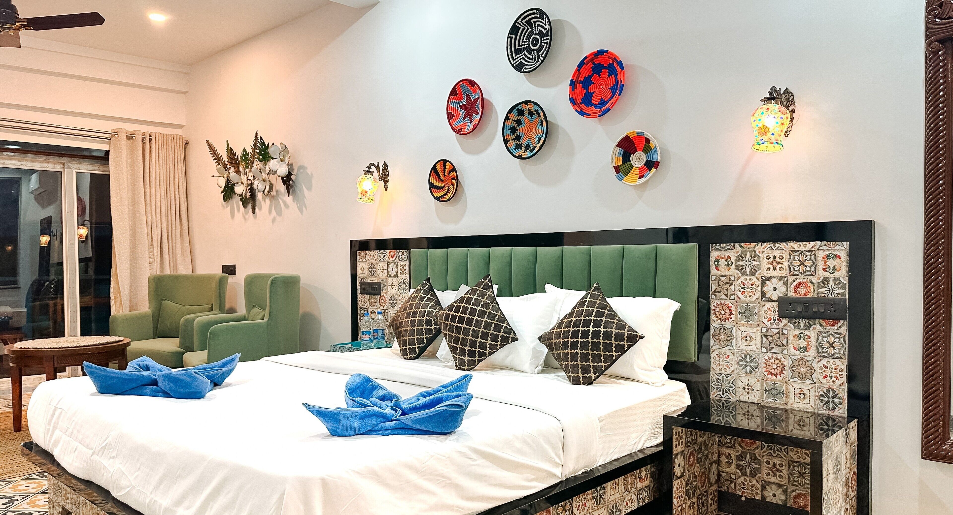Habitación de hotel con cabecera tapizada en verde, cama con ropa blanca y toallas azules, un sillón verde y decoración en la pared.