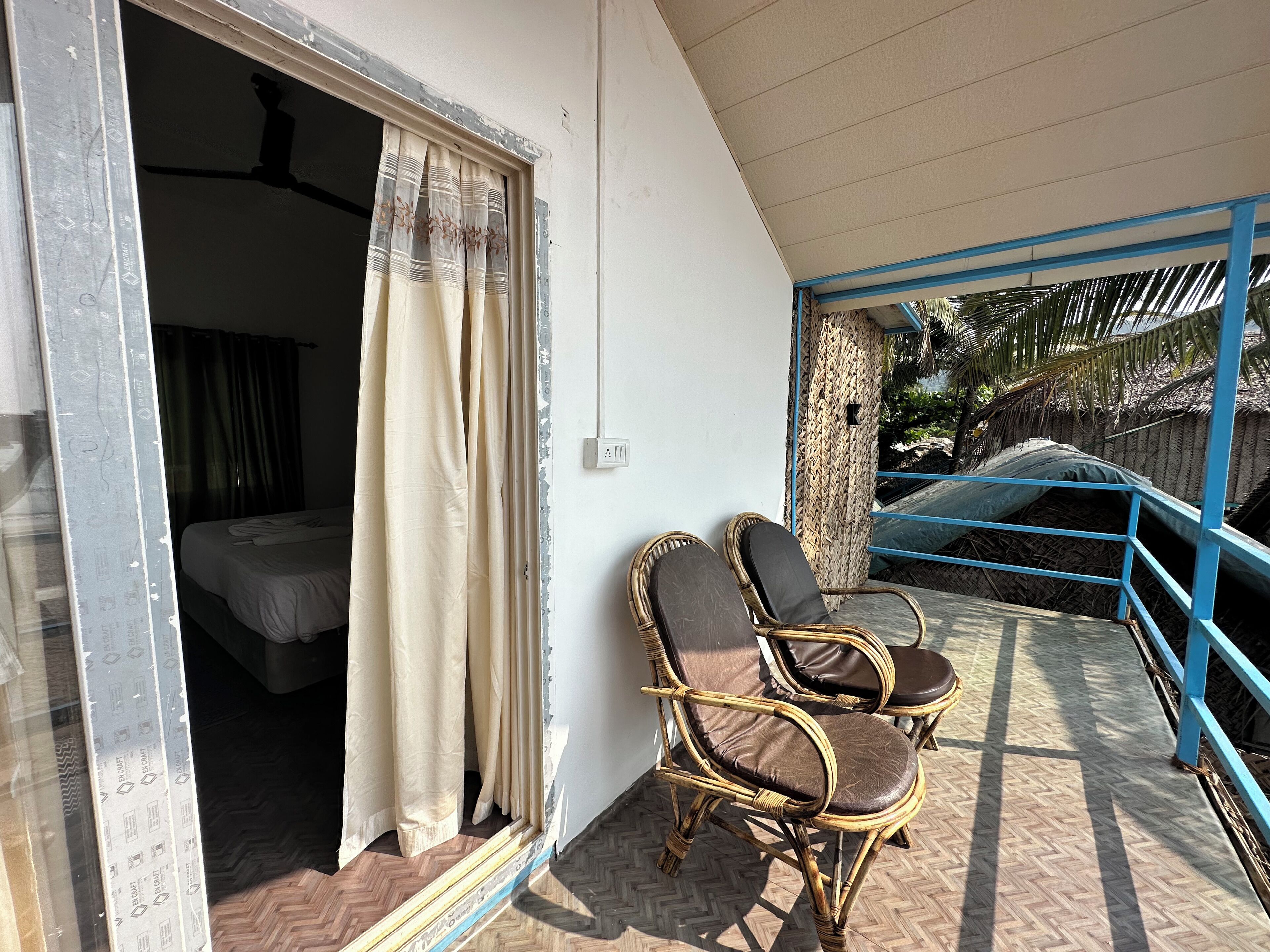A room with a bed, a window with curtains, and two wicker chairs on a balcony with a blue railing.