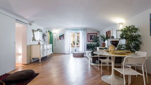 Un vaste espace de vie doté d’un plancher en bois, d’une table à manger avec des chaises, d’un meuble blanc et d’une grande fenêtre agrémentée de rideaux.