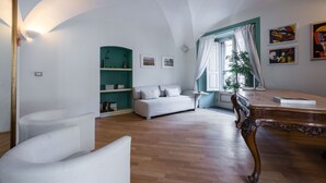 Un salon spacieux doté d’un parquet, d’un canapé blanc, d’un fauteuil blanc, d’une table basse en bois, d’une bibliothèque turquoise et d’une grande fenêtre ornée de rideaux blancs.