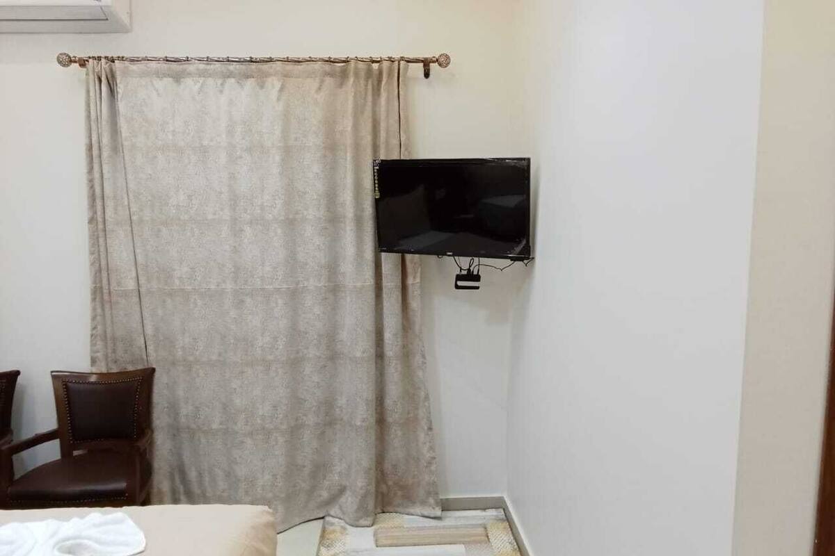 Une petite pièce simple, avec un lit, une chaise, un téléviseur fixé au mur et un rideau.