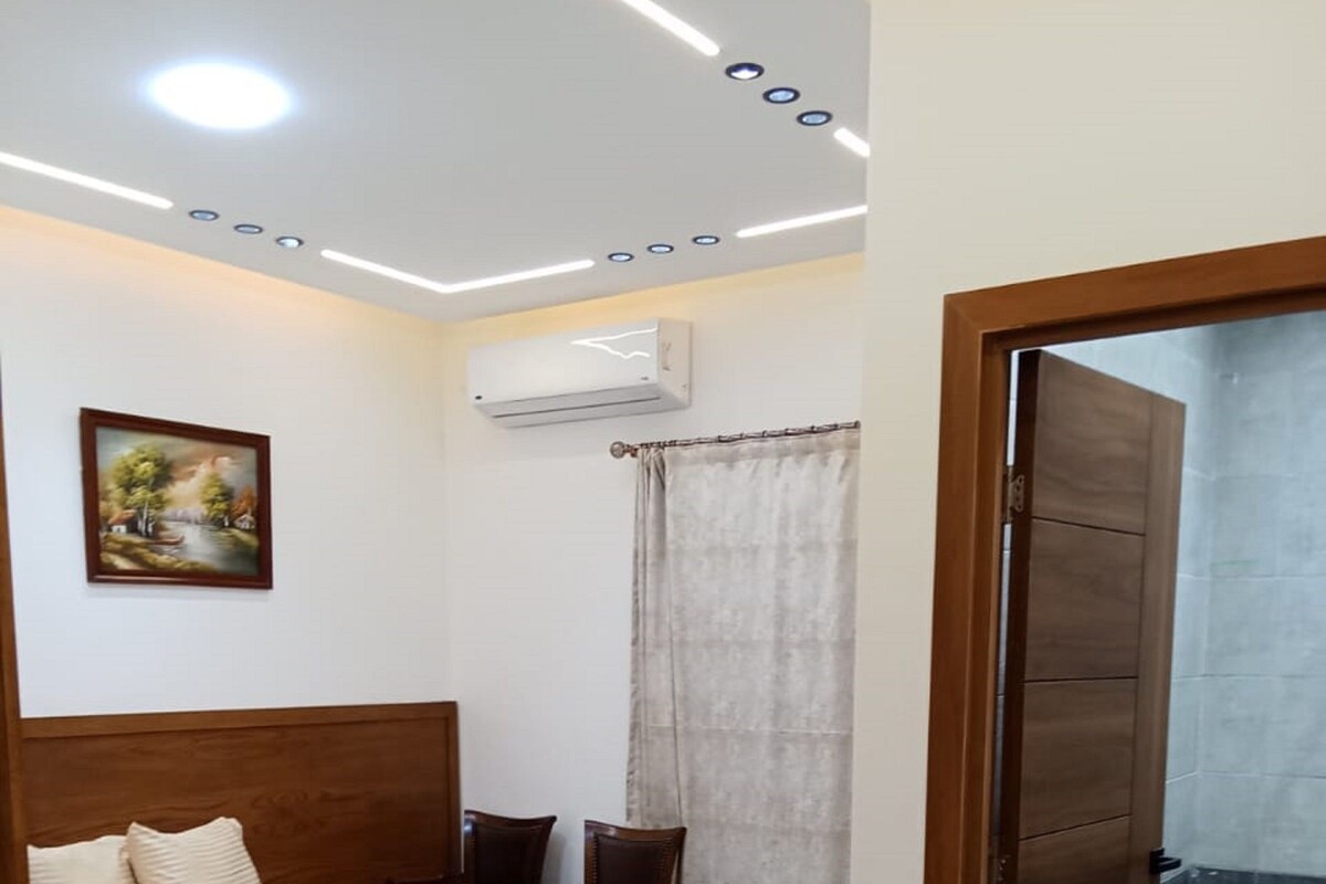 A bedroom with a bed, a chair, a desk, an air conditioner, a shower area, and a door.