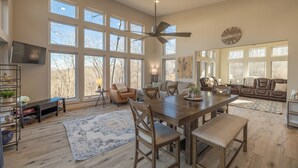 A spacious living room with a large wooden dining table, multiple chairs, a ceiling fan, and a large window with a view of trees. - Altitude Adjustment by Innsbrook Vacations! (Innsbrook)