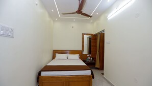Une chambre d’hôtel avec un grand lit, un ventilateur de plafond, des spots encastrés, une tête de lit en bois, une table de chevet avec une lampe et un miroir au mur.