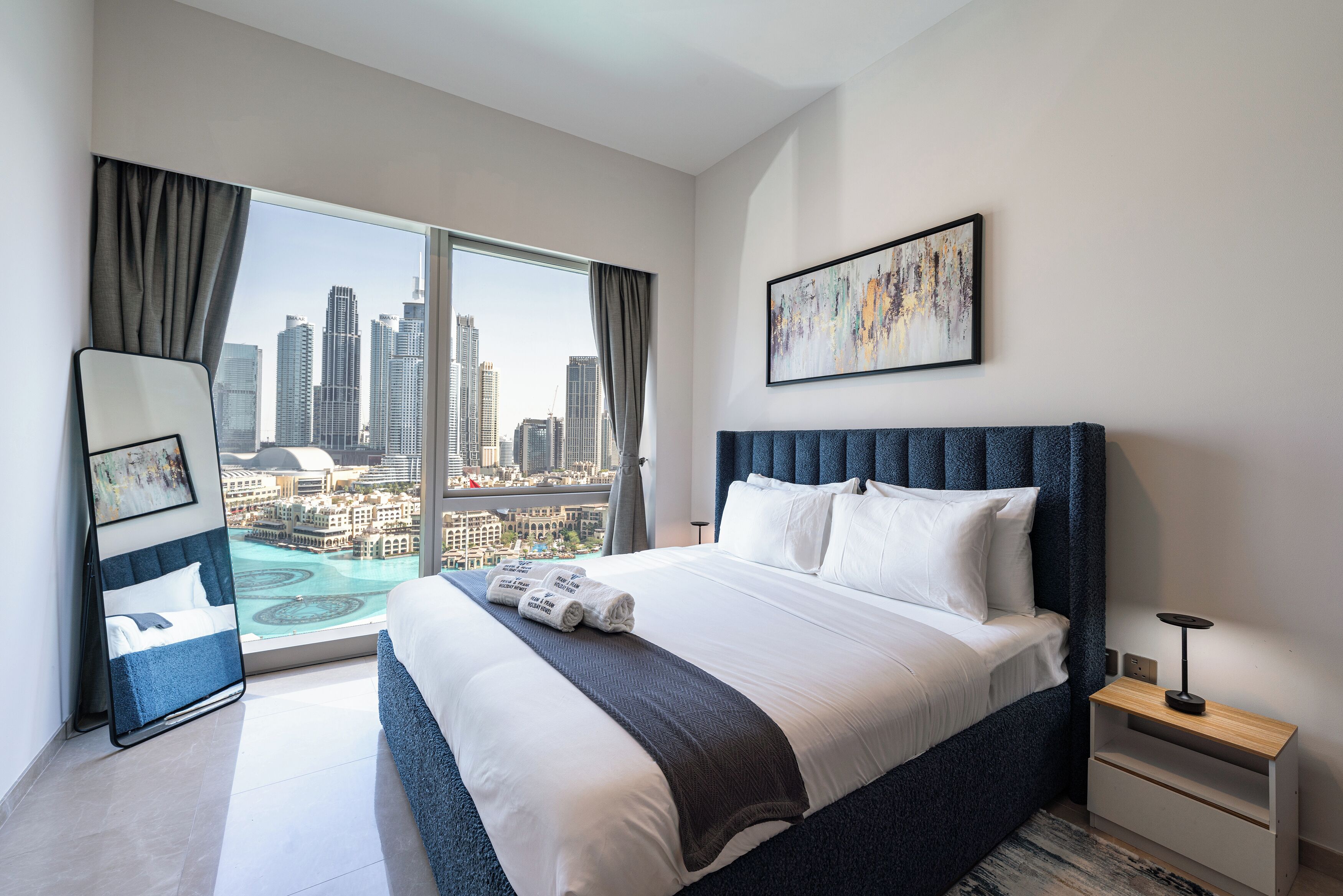 Ein modernes Hotelzimmer mit einem großen Bett, Blick auf die Stadt, einem gerahmten Kunstwerk und einem Nachttisch mit Lampe.