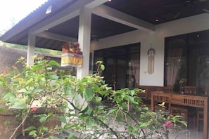 Un espace extérieur aménagé, abrité et doté d’une table et de chaises, entouré de verdure, avec une structure à toiture de chaume en arrière-plan.