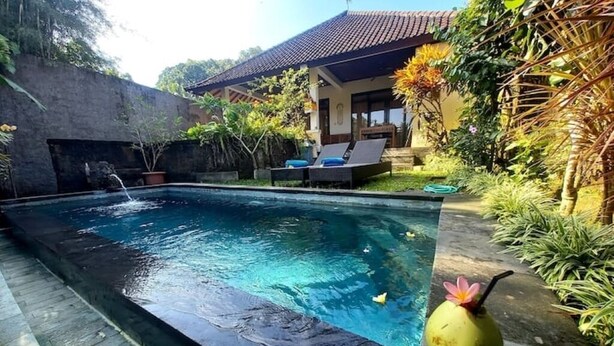 A rectangular pool with a stone edge, surrounded by lush greenery and a house with a tiled roof. - Private One BD with Pool Overlooking to Rice Fields By Hidden Point Villa

 (Bali)