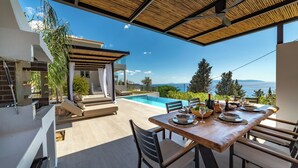 Zona pranzo a bordo piscina con vista sul mare, un'area salotto coperta e uno stile architettonico moderno.