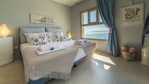 Un letto ordinato con cuscini decorati, una lampada da comodino e vista sul mare attraverso una finestra.