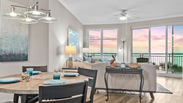 A modern dining area with a round table, chairs, and pendant lighting. There is a balcony with a view of the sunset. - Legacy Tower I Unit 302 | Overlooking the Beautiful Gulf! (Gulfport)