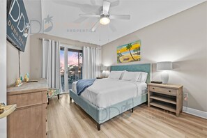 Une chambre à coucher avec un lit, des tables de chevet, un ventilateur de plafond, un tableau et une vue sur l’extérieur.