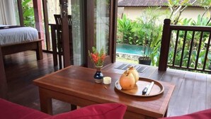 Uma mesa de madeira com um prato de comida, uma vela e um vaso de flores numa varanda com vista para uma piscina e vegetação.