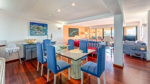 A dining area with a glass table and blue chairs, a living room with striped sofas, and artwork on the walls.