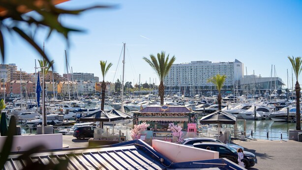 A marina with numerous boats docked, a building labeled "TIVOLI MARINA VILAMOURA" in the background, and a kiosk with pink flowers in the foreground.