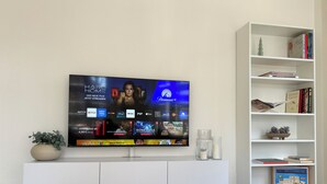 Moderni olohuone, jossa on valkoiselle seinälle asennettu litteänäyttöinen televisio, sen alla valkoinen viihdekeskus ja oikealla puolella kirjahylly.