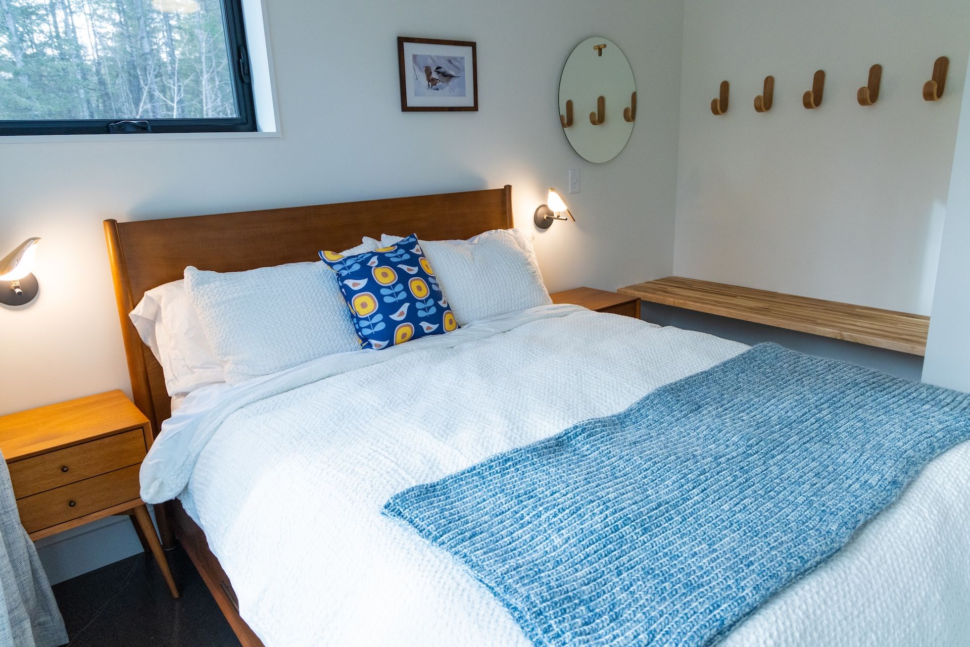 Un lit bien fait, avec une literie blanche et une couverture bleue, une tête de lit en bois, des tables de chevet avec des lampes, un tableau encadré au mur et un porte-manteau en bois.