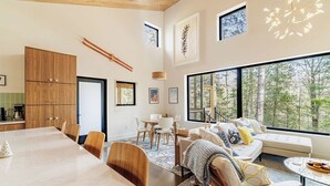 A modern interior with a dining area, a living space with a sofa and coffee table, and large windows offering a view of trees. - Cabin & Treehouse by Jay Cooke State Park / Duluth Trails (Esko)