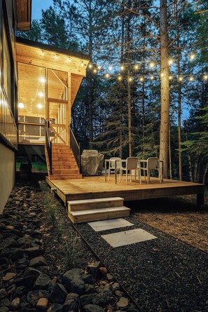 Terraza de madera con mobiliario exterior, una escalera que lleva a un nivel superior y un fondo de bosque.