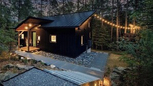 A small cabin with a dark exterior, a wooden deck, and a string of lights. - Cabin & Treehouse by Jay Cooke State Park / Duluth Trails (Esko)