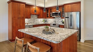 A kitchen with wooden cabinets, a stainless steel refrigerator, a built-in microwave, and a marble island with bar stools.