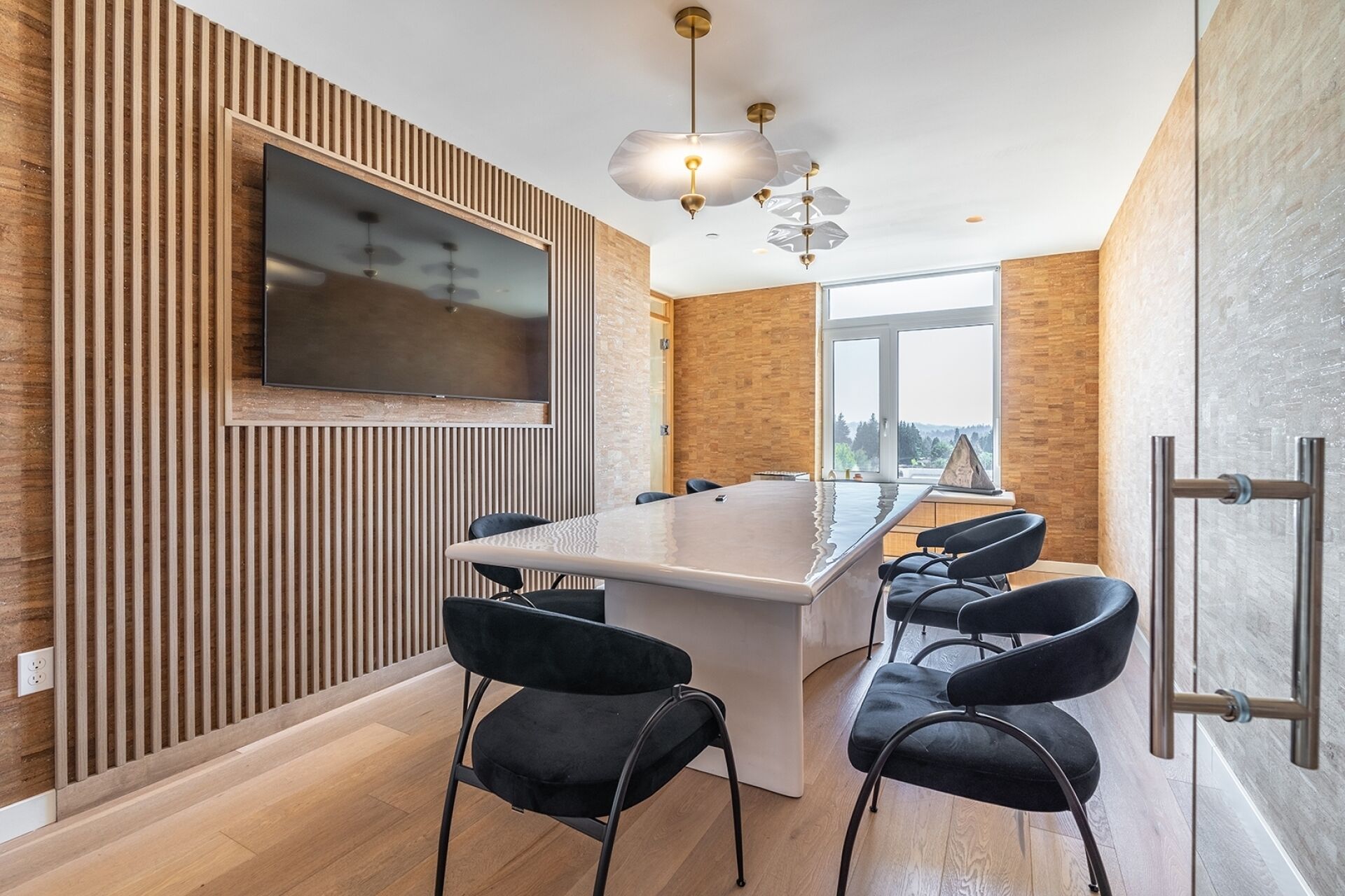 A modern kitchen with a central island, black chairs, a TV mounted on the wall, and a large window offering a view.