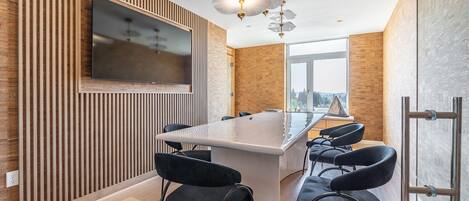 A modern kitchen with a central island, black chairs, a TV mounted on the wall, and a large window offering a view.